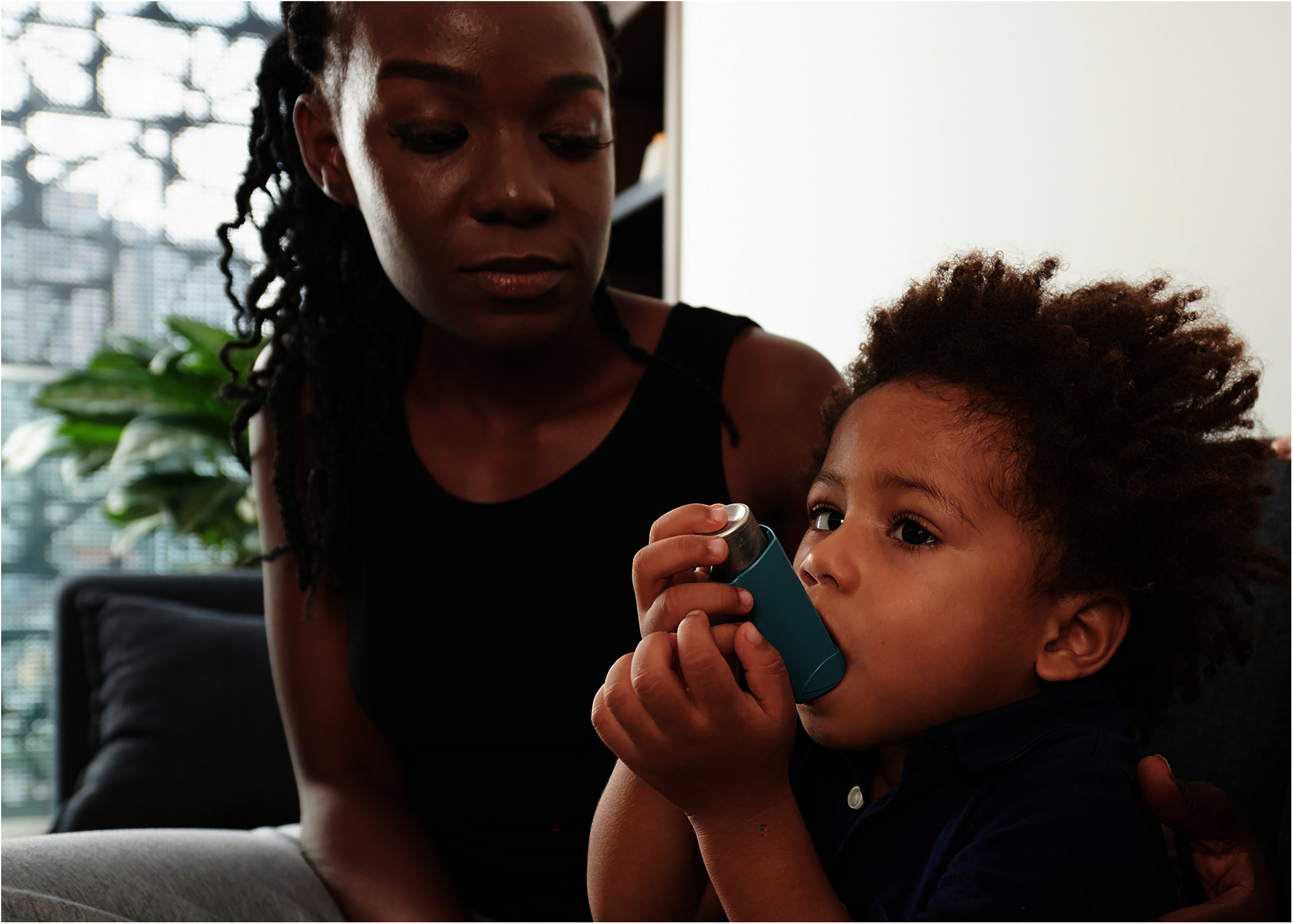 A concerned Black mother looks on as her young child uses an inhaler for asthma