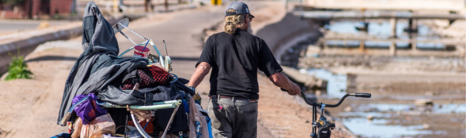 A man pushes a bicycle and a cart with belongings piled onto and hanging off of it, as he walks a path beside an empty, concrete-lined waterway