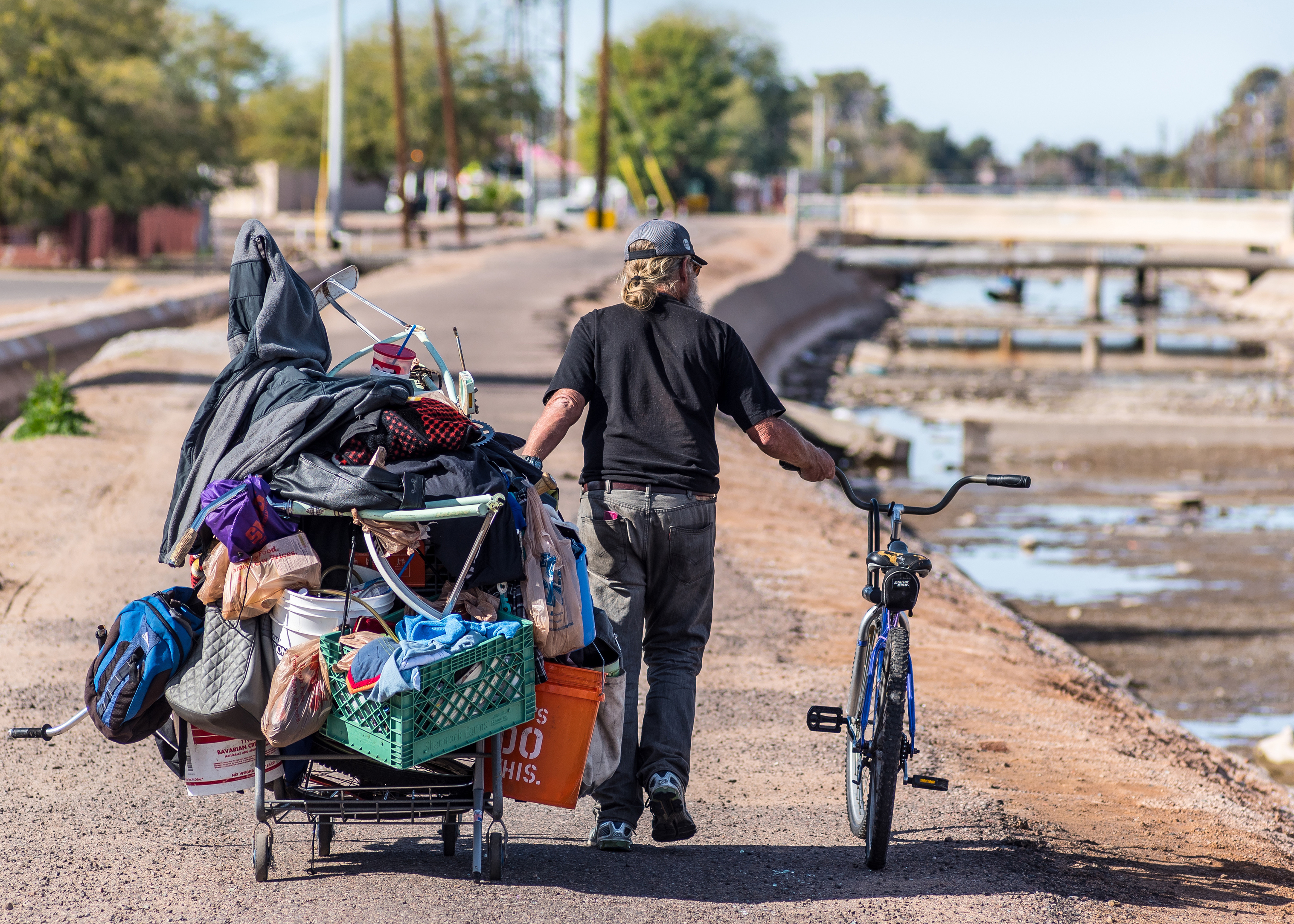 A man pushes a bicycle and a cart with belongings piled onto and hanging off of it, as he walks a path beside an empty, concrete-lined waterway