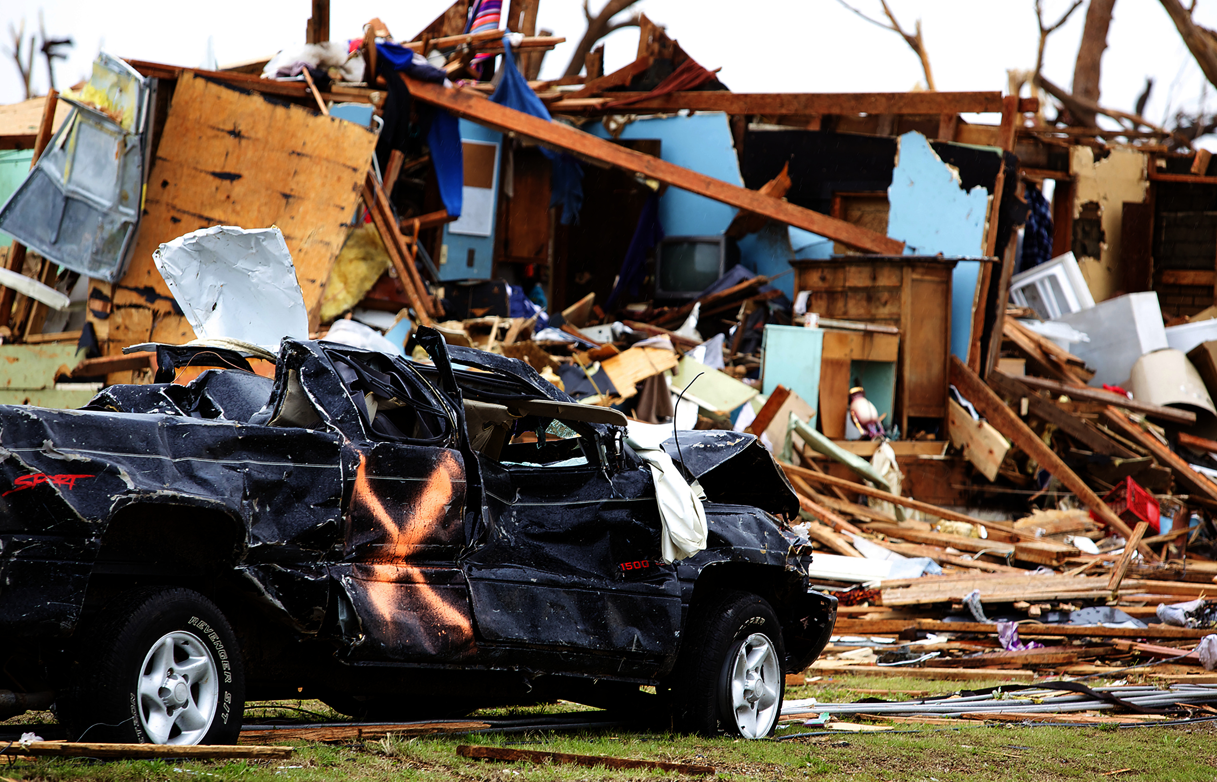 A dented, wrecked truck sits before a destroyed home, with jumbled fragments of wood, metal, and other construction material.