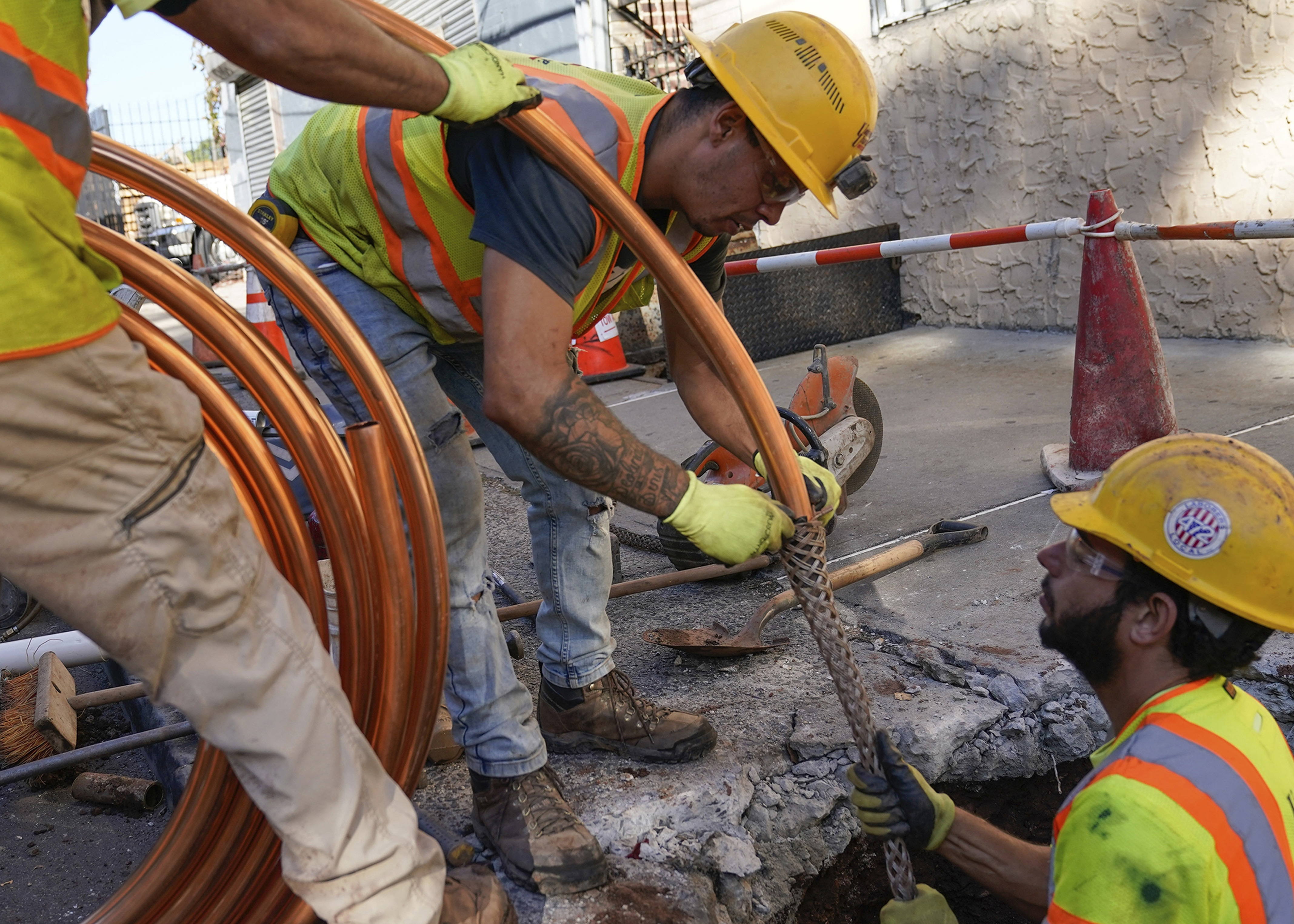 Construction worker holds large coil of copper pipe as another feeds it into a sleeve held by a third man standing in a hole dug into a city sidewalk