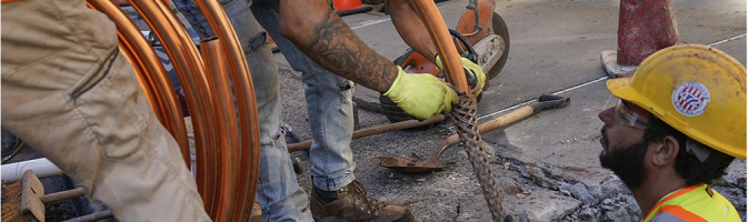 Construction worker holds large coil of copper pipe as another feeds it into a sleeve held by a third man standing in a hole dug into a city sidewalk