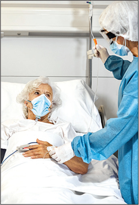 Female medical professional in protective suit adjusting IV drip of patient. Senior woman with oxygen mask is lying on bed in intensive care unit. They are in hospital during COVID-19 outbreak.