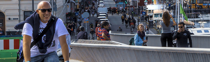 Middle-age man rides a bicycle over a canal lined with boats and homes on a sunny day
