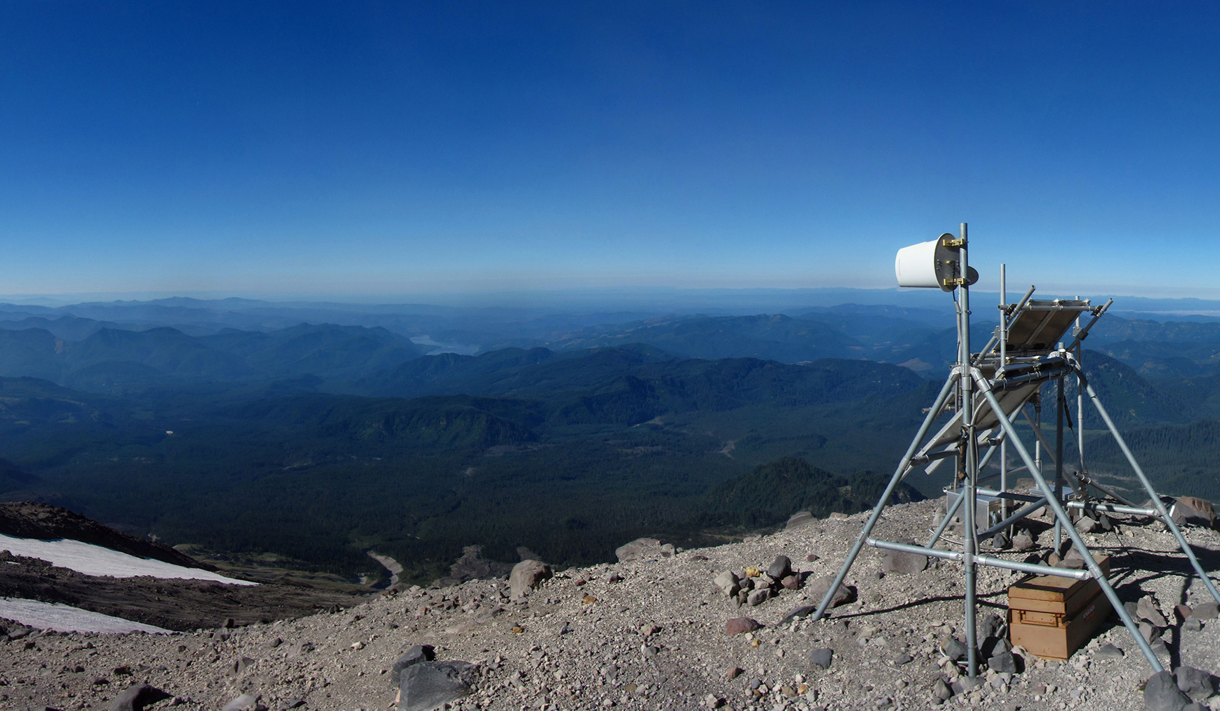 Monitoring device on the edge of Mount St. Helens, high above landscape with blue sky.