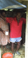 Figure 4: Fred stands in the foreground, holding onto a wooden bar above him to balance. His wife stands behind him, and is passing him a cup of water. There is a bowl in front of him.