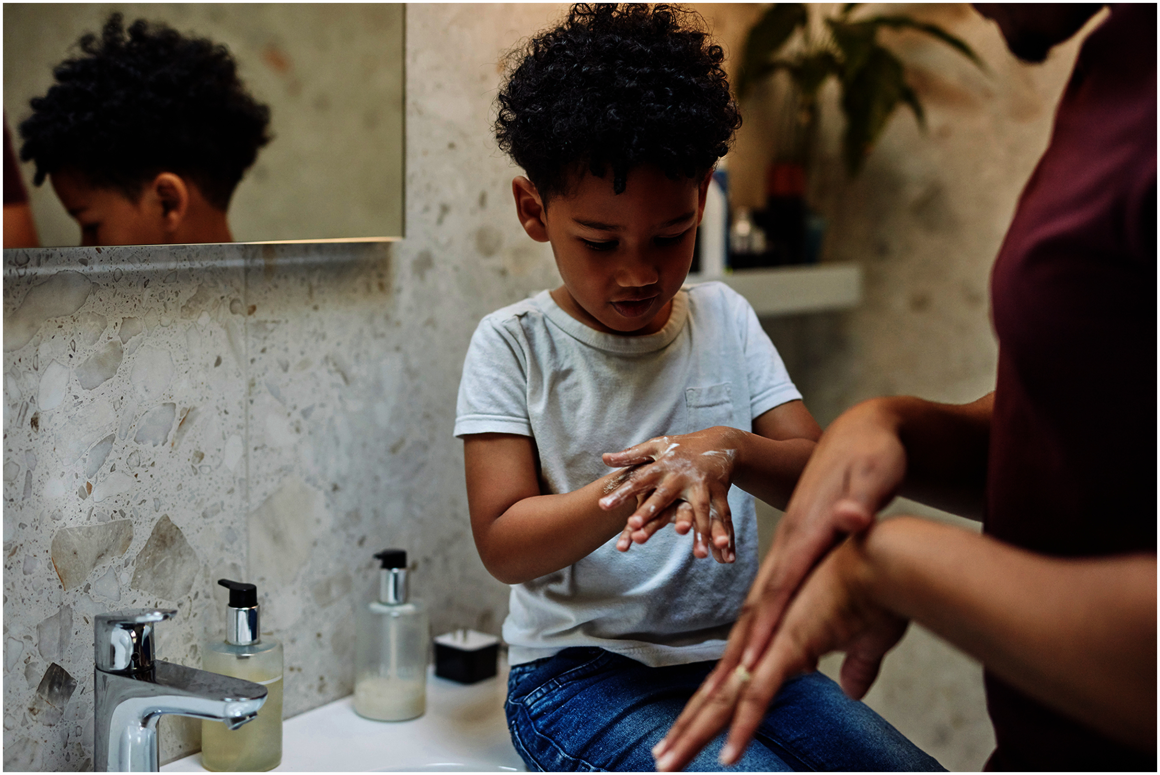 A child sitting on a bathroom sink rubs soap into his hands, as does an adult standing close by.