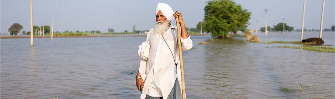 Man in a white turban, with a white beard, stands with a walking stick in a wide expanse of water that reaches above his ankles.