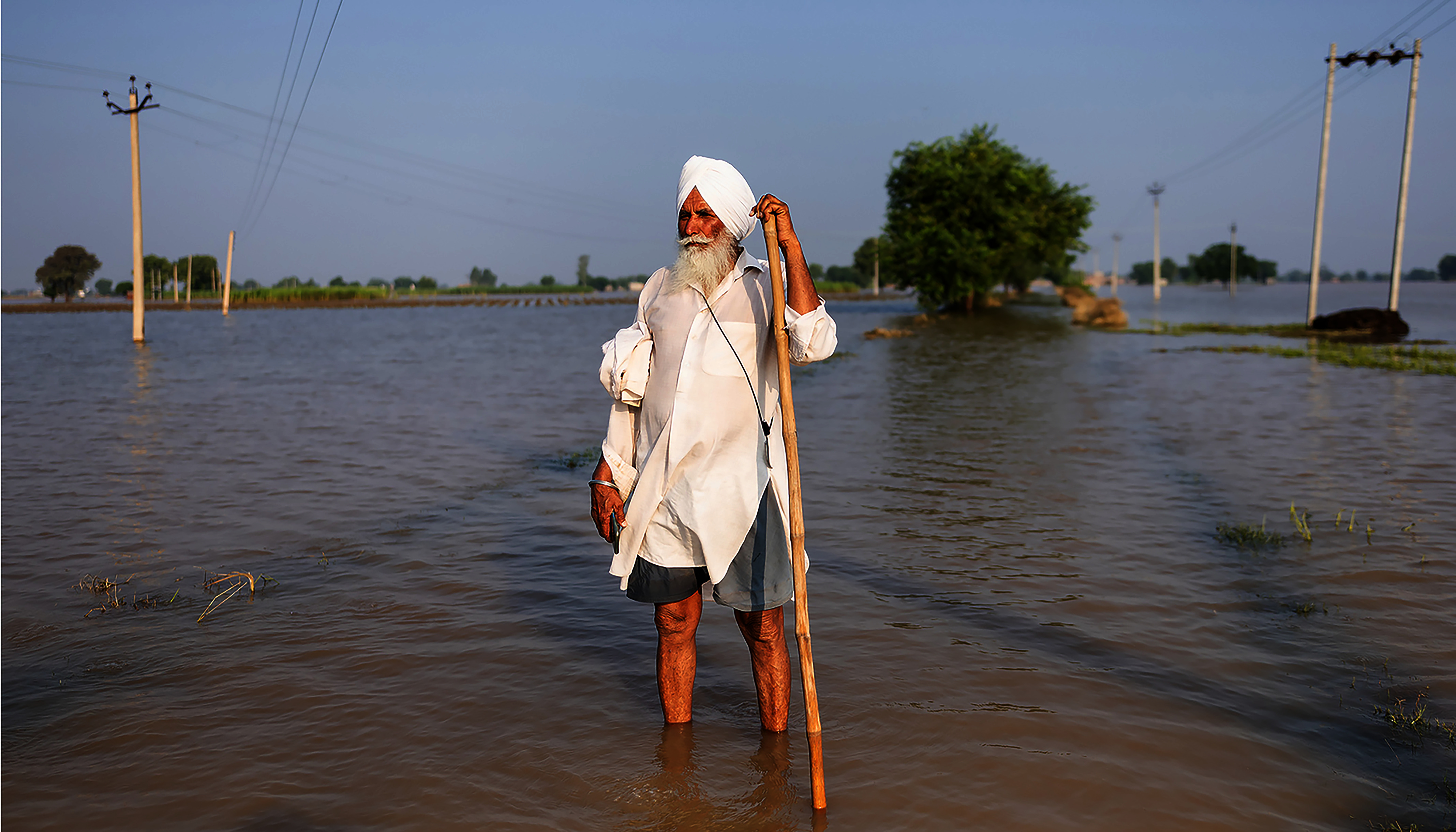 Man in a white turban, with a white beard, stands with a walking stick in a wide expanse of water that reaches above his ankles.