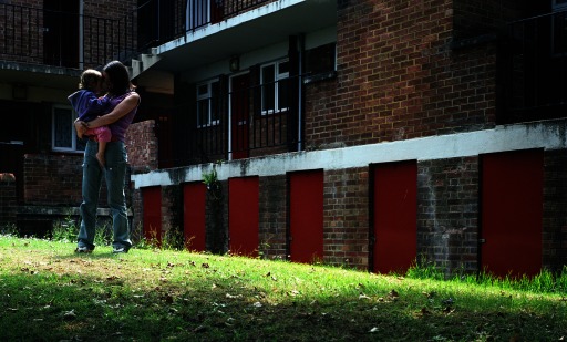 Photograph of a young mother holding her toddler daughter in front of subsidized housing.