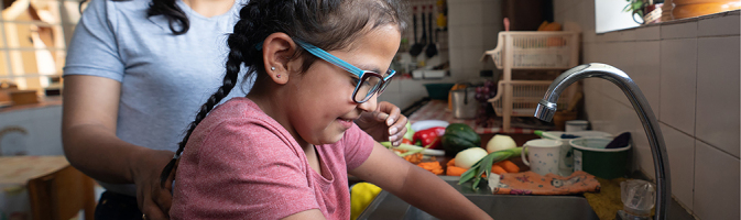 A Latin American girl washes vegetables at the kitchen sink with her mom close by