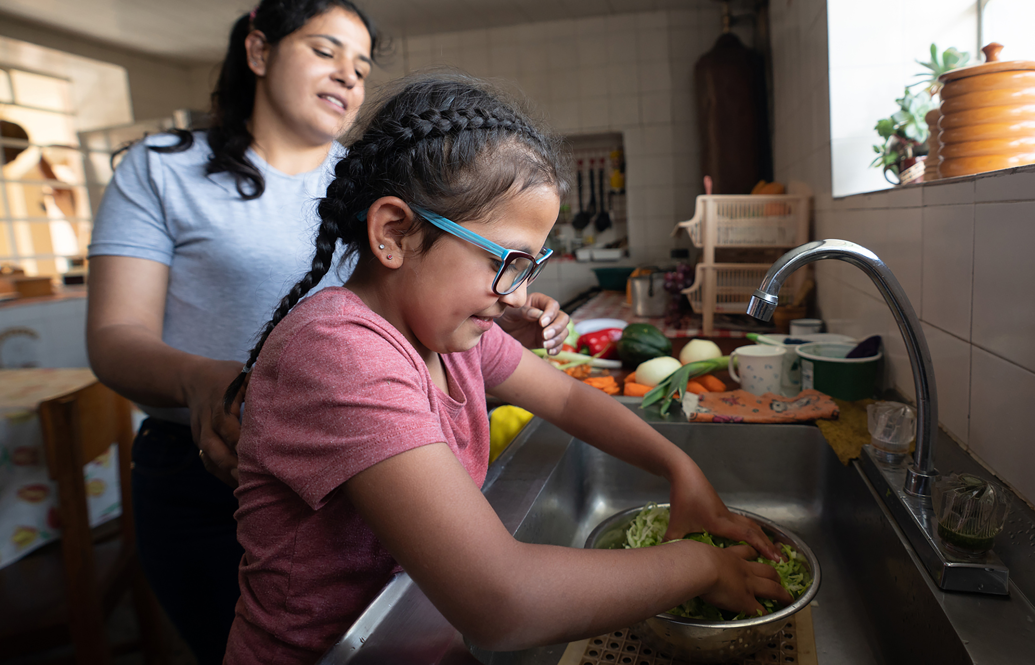 A Latin American girl washes vegetables at the kitchen sink with her mom close by