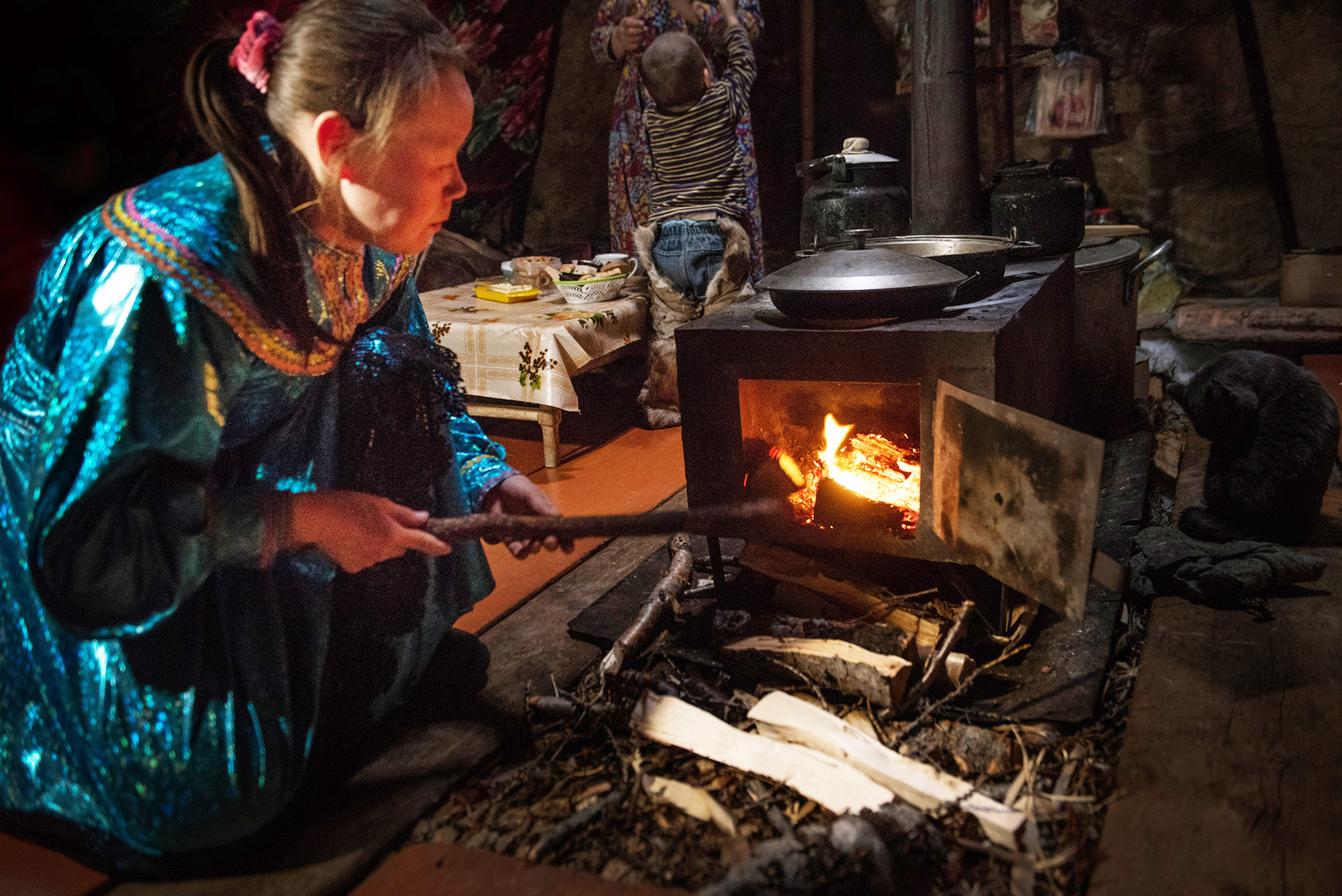 A woman stokes fire in small wood stove with cooking pans on top.