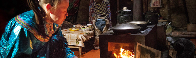 A woman stokes fire in small wood stove with cooking pans on top.